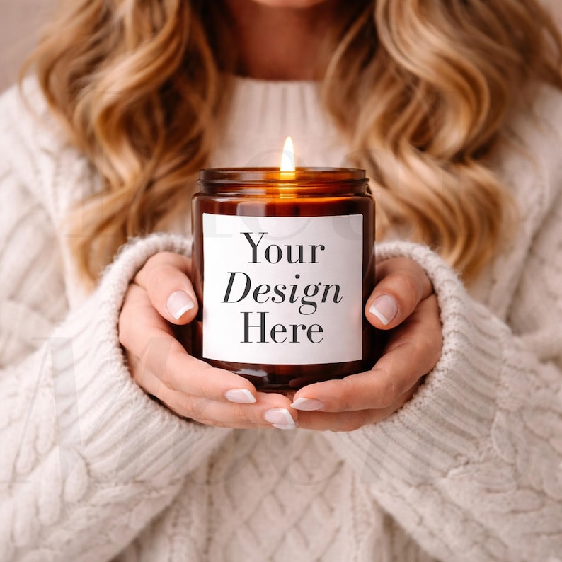 Op de afbeelding: Een brandende kaars in een bruine glazen pot met een wit etiket met de tekst "Your Design Here". De kaars wordt met beide handen vastgehouden. De achtergrond is wazig en toont een persoon die een witte trui draagt.
