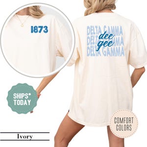 May include: Ivory-colored t-shirt with blue text and graphics. The back of the shirt features the words "DELTA GAMMA" in a stacked design, with "dee gee" in a lighter blue. The front has the number "1873" in blue.