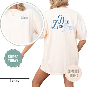 May include: Ivory-colored t-shirt with blue text and bow design. The front of the shirt has a small bow graphic and the text "Est 1902". The back features the name "Dee Lee" in script with a bow graphic. The shirt is a comfort color.