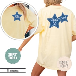 May include: A pale yellow short-sleeve t-shirt featuring a blue star graphic on the front left chest with Greek letters. The back displays two blue stars with the text Alpha Xi and 1893. The design has a distressed texture throughout the blue stars.
