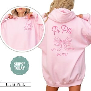 May include: Light pink hooded sweatshirt with the text "Pi Phi Est. 1867" and a pink bow on the back. The front of the sweatshirt has a small pink bow with the text "Est. 1867".