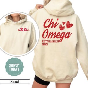 May include: Sand-colored hoodie with red text and heart graphics. The front features the Greek letters ΧΩ with heart accents. The back displays "Chi Omega" with "Established 1895" below. The hoodie has a hood and long sleeves.