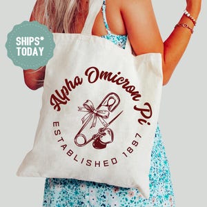 May include: Cream-colored tote bag with a maroon graphic of a safety pin, bow, and heart, with the text "Alpha Omicron Pi ESTABLISHED 1897". The bag has a light-colored handle.