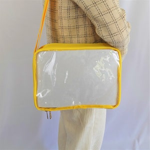 K&ouml;nnte beinhalten: Eine gelbe transparente Tragetasche mit Rei&szlig;verschluss und Schultergurt. Die Tasche ist aus strapazierf&auml;higem Stoff gefertigt und hat ein transparentes Plastikfenster auf der Vorderseite.
