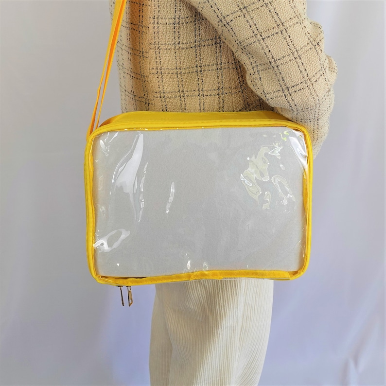 K&ouml;nnte beinhalten: Eine gelbe transparente Tragetasche mit Rei&szlig;verschluss und Schultergurt. Die Tasche ist aus strapazierf&auml;higem Stoff gefertigt und hat ein transparentes Plastikfenster auf der Vorderseite.