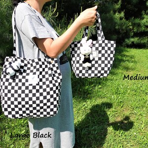 K&ouml;nnte beinhalten: Zwei schwarz-wei&szlig; karierte Tragetaschen, eine gro&szlig;e und eine mittlere, werden gezeigt. An jeder Tasche ist ein Stofftier befestigt. Die Taschen werden vor einem gr&uuml;nen Hintergrund gehalten. Der Text "Large Black" und "Medium Black" ist sichtbar.