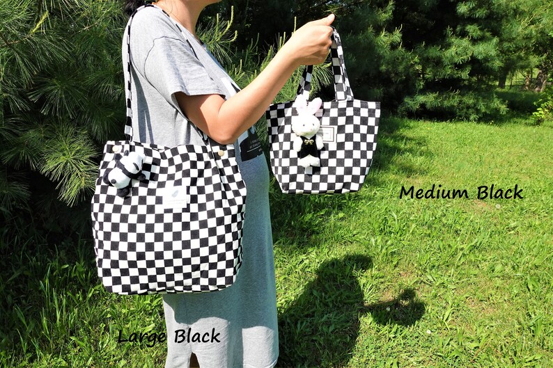 K&ouml;nnte beinhalten: Zwei schwarz-wei&szlig; karierte Tragetaschen, eine gro&szlig;e und eine mittlere, werden gezeigt. An jeder Tasche ist ein Stofftier befestigt. Die Taschen werden vor einem gr&uuml;nen Hintergrund gehalten. Der Text "Large Black" und "Medium Black" ist sichtbar.