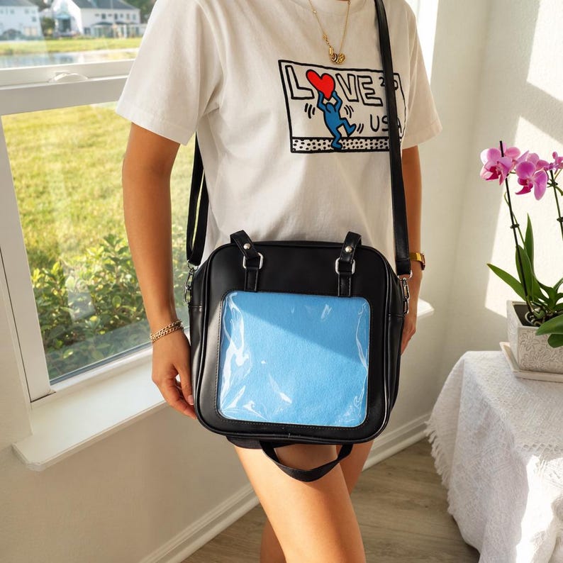 Puede incluir: Bolso bandolera cuadrado negro con un bolsillo frontal transparente que contiene un inserto azul claro. El bolso tiene un asa superior y una correa larga para el hombro. La persona lleva una camiseta blanca con la palabra "LOVE" y un gr&aacute;fico.