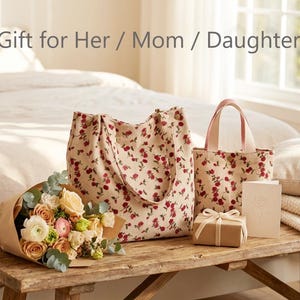 May include: A still-life arrangement featuring a bouquet of flowers, two tote bags, a gift box, and a card on a wooden bench. The bags and bouquet have a floral pattern. The text "Gift for Her / Mom / Daughter" is at the top.