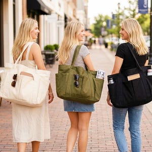 May include: Three women walking down a brick-paved street, each carrying a large tote bag. The bags are cream, olive green, and black. Each bag has multiple pockets and compartments. Sunglasses are visible on the bags.