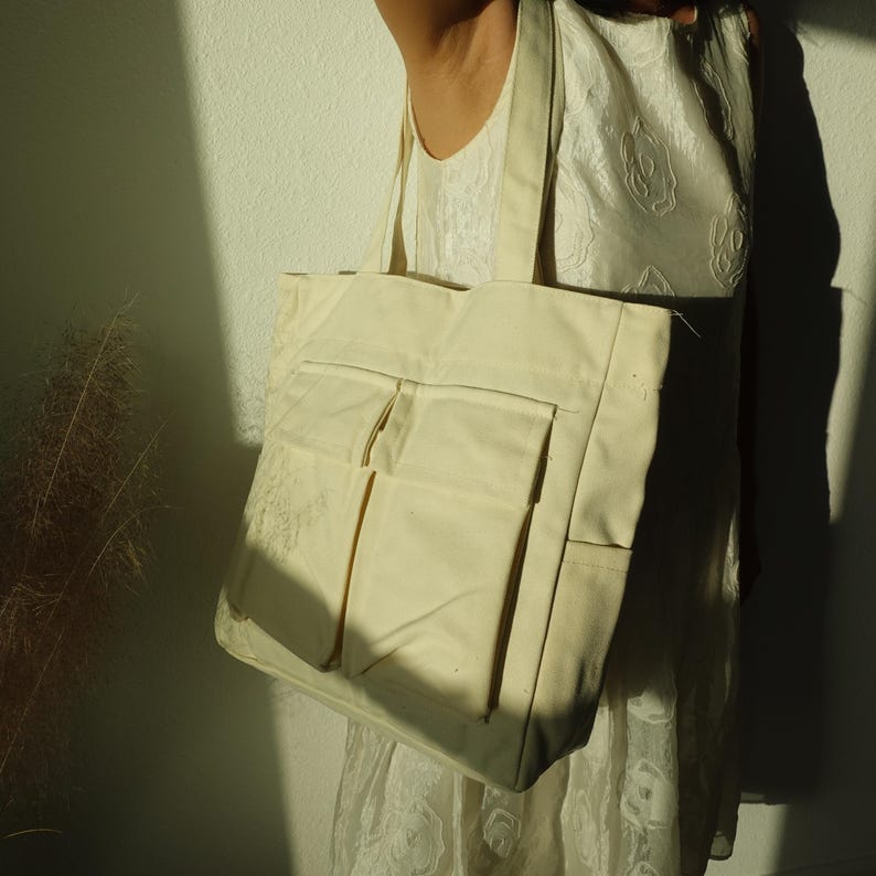 Pode incluir: Bolsa tote de lona creme com dois bolsos frontais. A bolsa tem formato retangular e al&ccedil;as longas para carregar no ombro. A bolsa &eacute; feita de um material resistente.