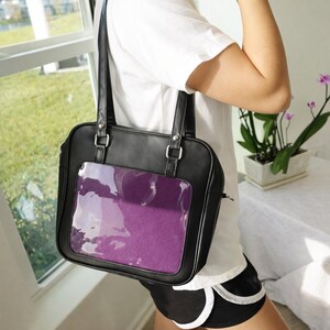 Puede incluir: Bolso tote de cuero sint&eacute;tico negro con un bolsillo frontal transparente que muestra un inserto morado. El bolso tiene dos asas y herrajes plateados. El bolso es de forma cuadrada y tiene cierre de cremallera.