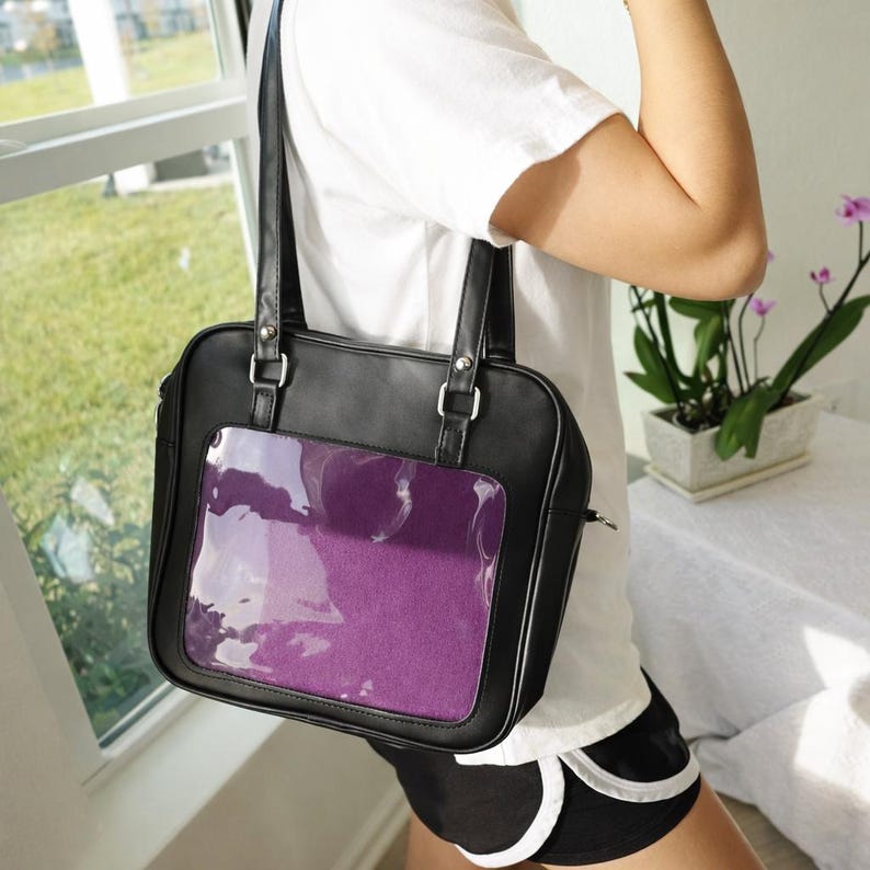 Puede incluir: Bolso tote de cuero sint&eacute;tico negro con un bolsillo frontal transparente que muestra un inserto morado. El bolso tiene dos asas y herrajes plateados. El bolso es de forma cuadrada y tiene cierre de cremallera.