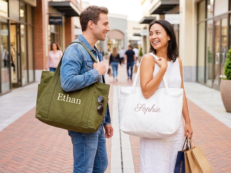 May include: A couple walking in an outdoor shopping area, each carrying a large tote bag. One bag is olive green with the name "Ethan" in white, and the other is white with "Sophie" in a script font. Both bags have pockets.