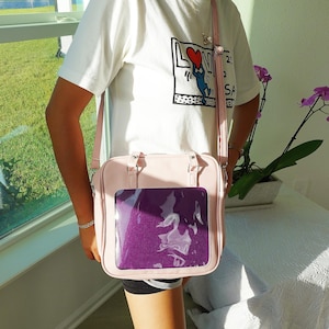 Puede incluir: Un bolso de hombro rosa claro con una ventana frontal transparente que muestra un inserto morado. El bolso tiene un asa superior y una correa ajustable. La persona lleva una camiseta blanca con un estampado gr&aacute;fico.