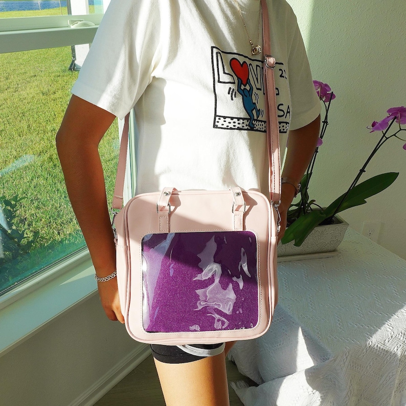 Puede incluir: Un bolso de hombro rosa claro con una ventana frontal transparente que muestra un inserto morado. El bolso tiene un asa superior y una correa ajustable. La persona lleva una camiseta blanca con un estampado gr&aacute;fico.