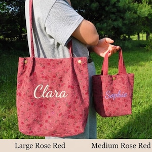 May include: Two rose red corduroy tote bags, one large and one medium, with floral patterns. The large bag has "Clara" embroidered in white, and the smaller bag has "Sophie" embroidered in purple. Both bags have handles and are held by a person.