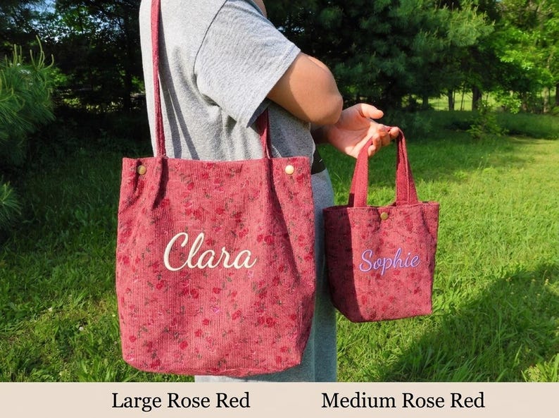 May include: Two rose red corduroy tote bags, one large and one medium, with floral patterns. The large bag has "Clara" embroidered in white, and the smaller bag has "Sophie" embroidered in purple. Both bags have handles and are held by a person.