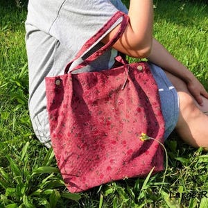 May include: A red corduroy tote bag with a floral pattern. The bag has two handles and is secured with two buttons. The bag is sitting on a person's lap in a grassy area.