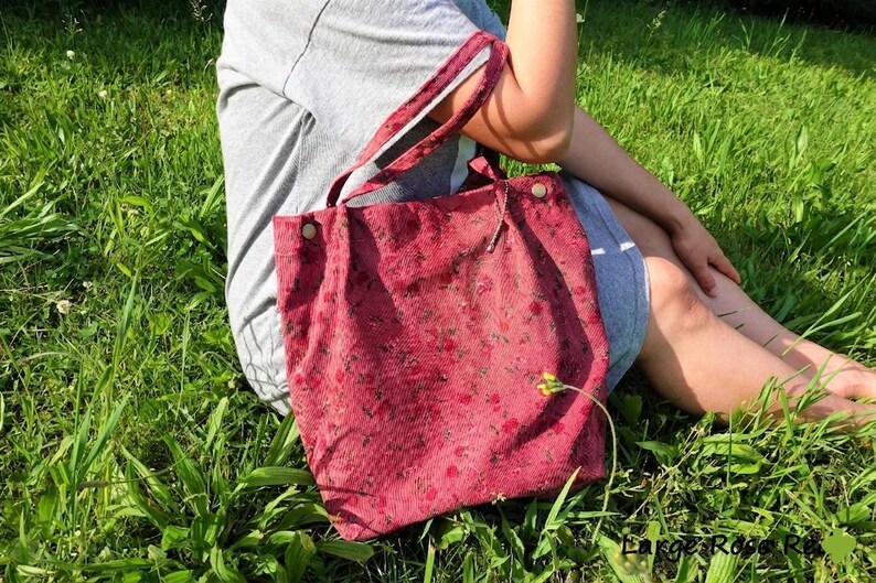 May include: A red corduroy tote bag with a floral pattern. The bag has two handles and is secured with two buttons. The bag is sitting on a person's lap in a grassy area.