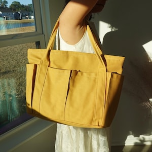 May include: Mustard yellow tote bag with multiple exterior pockets and sturdy handles. The bag is made of a durable canvas-like material. The tote bag is held in front of a window with a view of a lake and houses.