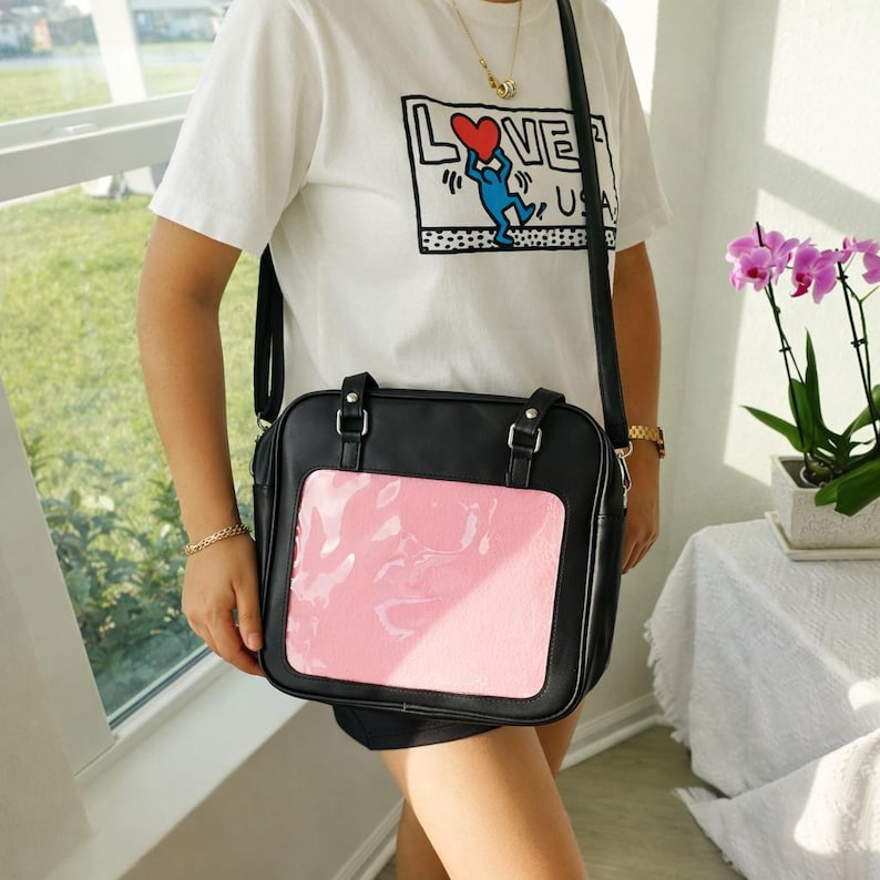 Puede incluir: Bolso bandolera negro con un bolsillo frontal rosa transparente. El bolso tiene forma rectangular con un asa superior y una correa larga para el hombro. La persona lleva una camiseta blanca con un estampado gr&aacute;fico.