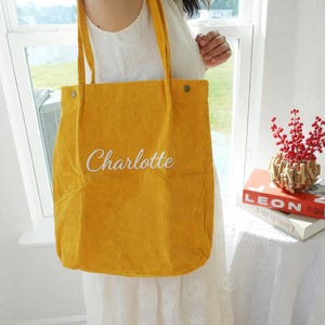 May include: Mustard yellow corduroy tote bag with the name "Charlotte" embroidered in white script. The bag has long straps and is held up by a person. The bag is a simple, casual style.