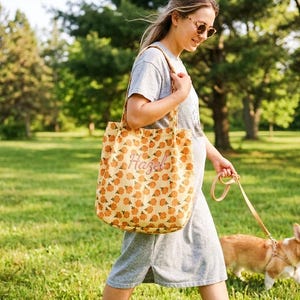 May include: A beige tote bag with a repeating pattern of orange pumpkins and the name "Hazel" embroidered on the front. The bag is carried by a person walking a dog in a park. The dog is a corgi.