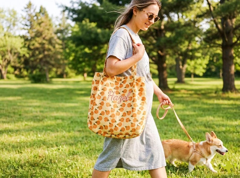 May include: A beige tote bag with a repeating pattern of orange pumpkins and the name "Hazel" embroidered on the front. The bag is carried by a person walking a dog in a park. The dog is a corgi.