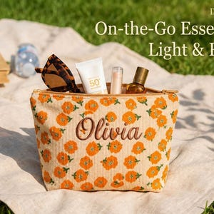 May include: A cream corduroy cosmetic bag with an orange floral pattern and the name "Olivia" embroidered on the front. The bag contains sunglasses, sunscreen, and other beauty products. The text "On-the-Go Essentials Light & Ready" is visible.