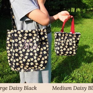 May include: Two black daisy-patterned tote bags, one large and one medium, are displayed against a green grassy background. The large bag has a shoulder strap, and both bags feature embroidered names. The medium bag has red handles.