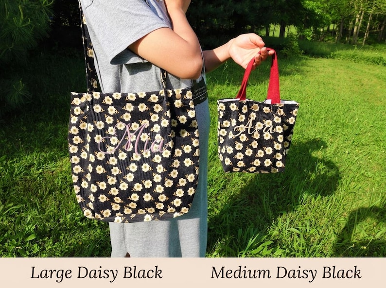 May include: Two black daisy-patterned tote bags, one large and one medium, are displayed against a green grassy background. The large bag has a shoulder strap, and both bags feature embroidered names. The medium bag has red handles.