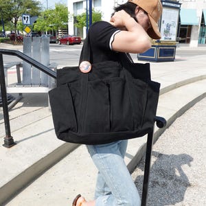 May include: A large black tote bag with multiple exterior pockets. The bag is made of a sturdy fabric and features a circular decorative pin. The bag is being carried outdoors on a sunny day.