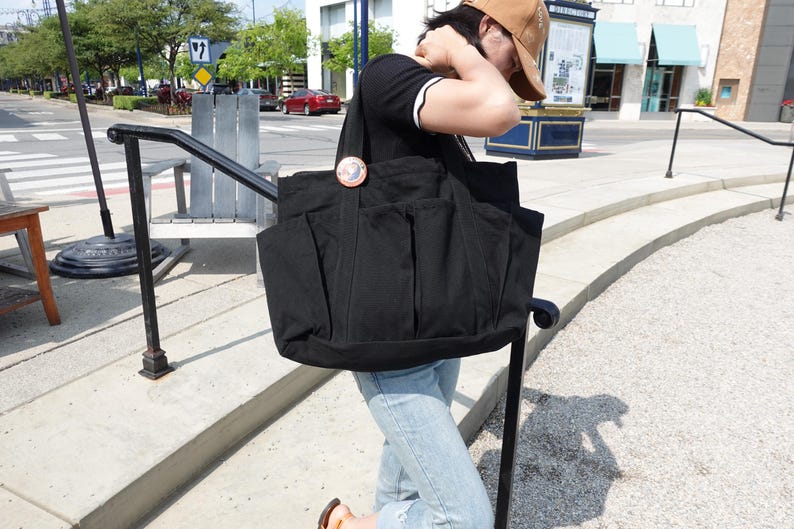 May include: A large black tote bag with multiple exterior pockets. The bag is made of a sturdy fabric and features a circular decorative pin. The bag is being carried outdoors on a sunny day.