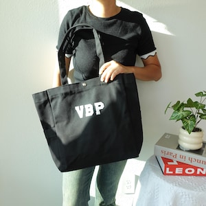 Może przedstawiać: Czarna torba typu tote z białymi literami "VBP". Torba ma dwa uchwyty i zapięcie na zatrzask. Torba jest wykonana z wytrzymałego materiału i ma dwie boczne kieszenie. Prosta torba na co dzień.