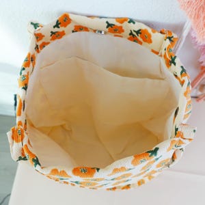 May include: Open view of a cream-colored corduroy tote bag with a floral pattern in orange and green. The interior is a solid light beige color. The bag has a drawstring closure and a small pocket inside.