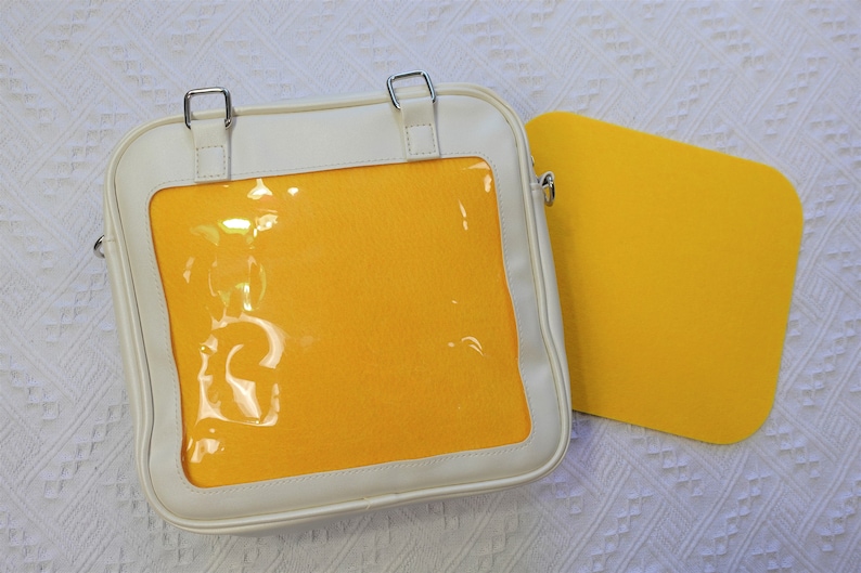 K&ouml;nnte beinhalten: Eine wei&szlig;e Tasche aus Kunstleder mit einem durchsichtigen Plastikfenster, das ein gelbes Innenleben zeigt. Die Tasche hat einen Schultergurt und ein kleines gelbes Quadrat, das an ihr befestigt ist.