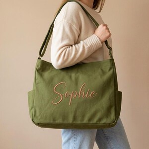 May include: Olive green canvas tote bag with the name "Sophie" embroidered in a cursive font. The bag has two exterior pockets and a shoulder strap. The person is wearing a cream-colored sweater and blue jeans.