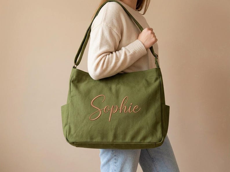 May include: Olive green canvas tote bag with the name "Sophie" embroidered in a cursive font. The bag has two exterior pockets and a shoulder strap. The person is wearing a cream-colored sweater and blue jeans.