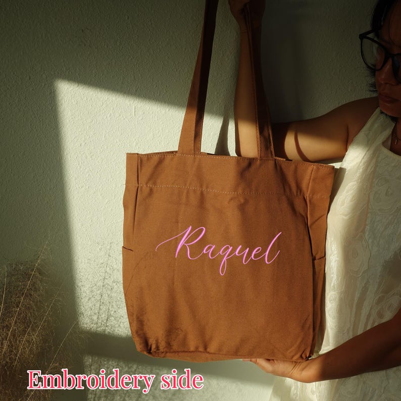 Pode incluir: Uma sacola de lona marrom com o nome "Raquel" bordado em rosa. A sacola tem duas al&ccedil;as e formato retangular. As palavras "Embroidery side" est&atilde;o na parte inferior da imagem.