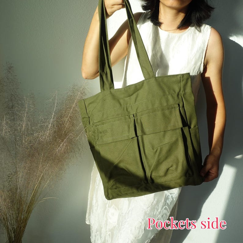 Pode incluir: Bolsa tote verde-oliva com dois bolsos frontais e al&ccedil;as longas. A bolsa &eacute; feita de um tecido resistente. O texto "Pockets side" &eacute; vis&iacute;vel no canto inferior direito da imagem. A bolsa est&aacute; sendo segurada por uma pessoa.