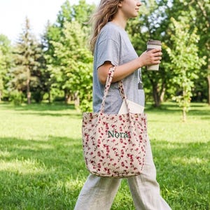 May include: A floral print tote bag with the name "Nora" embroidered in green. The bag has long straps and is filled with a white tote bag. The person is holding a reusable coffee cup. The background is a green park.
