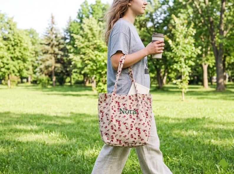 May include: A floral print tote bag with the name "Nora" embroidered in green. The bag has long straps and is filled with a white tote bag. The person is holding a reusable coffee cup. The background is a green park.