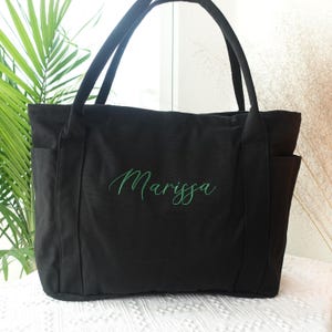 May include: Black tote bag with two exterior pockets and long handles. The name "Marissa" is embroidered in green script across the front. The bag is made of a sturdy fabric and is suitable for carrying various items.