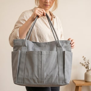Op de afbeelding: Een grote, grijze tote bag met meerdere buitenvakken en lange handvatten. De tas is gemaakt van een stevige stof en wordt vastgehouden. De tote bag is een praktische accessoire voor het dragen van spullen.