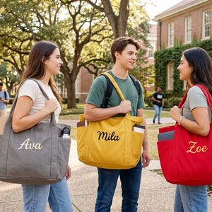 May include: Three students stand on a college campus, each carrying a personalized tote bag. The bags are gray, yellow, and red, with the names "Ava", "Mila", and "Zoe" embroidered on them. The scene suggests a back-to-school theme.