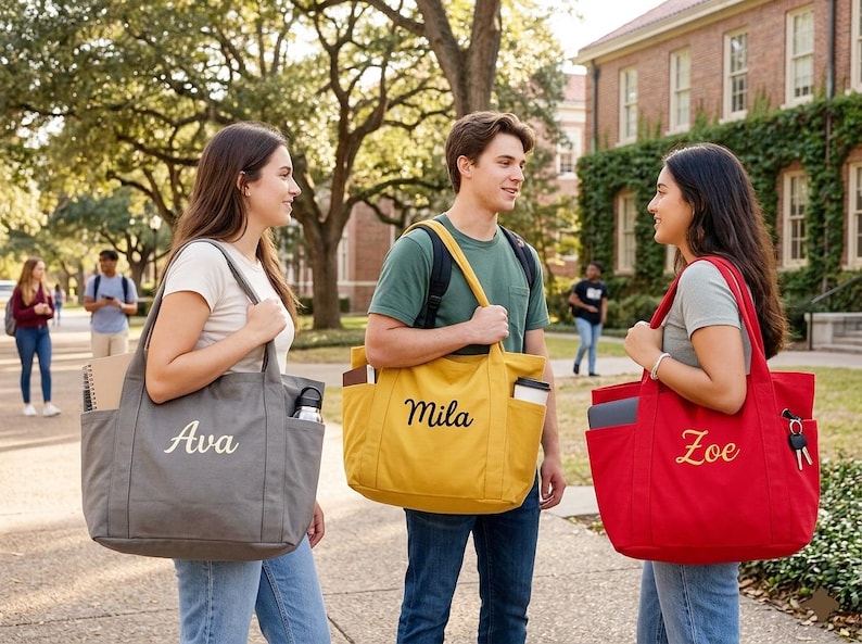 May include: Three students stand on a college campus, each carrying a personalized tote bag. The bags are gray, yellow, and red, with the names "Ava", "Mila", and "Zoe" embroidered on them. The scene suggests a back-to-school theme.