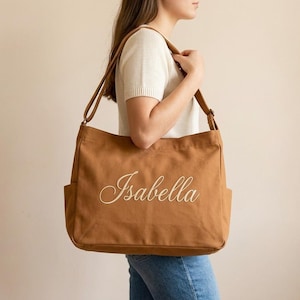 May include: A brown canvas tote bag with the name "Isabella" embroidered in a cursive font. The bag has two exterior pockets and a long shoulder strap. The bag is carried over the shoulder.