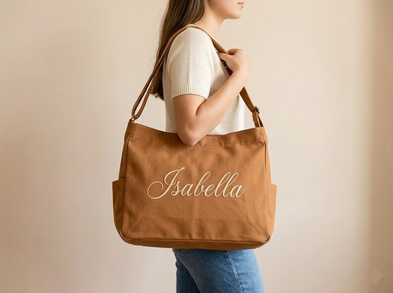May include: A brown canvas tote bag with the name "Isabella" embroidered in a cursive font. The bag has two exterior pockets and a long shoulder strap. The bag is carried over the shoulder.