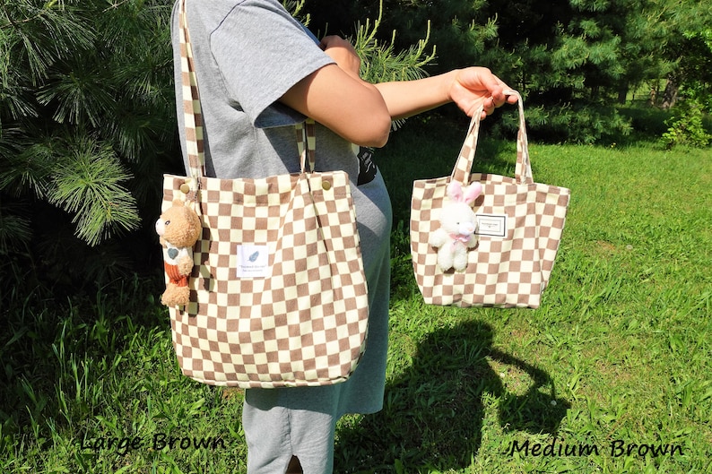 K&ouml;nnte beinhalten: Zwei braun-wei&szlig; karierte Tragetaschen, eine gro&szlig;e und eine mittlere. Die gro&szlig;e Tasche hat einen Schultergurt und einen Teddyb&auml;r-Schl&uuml;sselanh&auml;nger. Die mittlere Tasche hat einen wei&szlig;en Hasen-Schl&uuml;sselanh&auml;nger. Beide Taschen haben ein kleines Etikett.